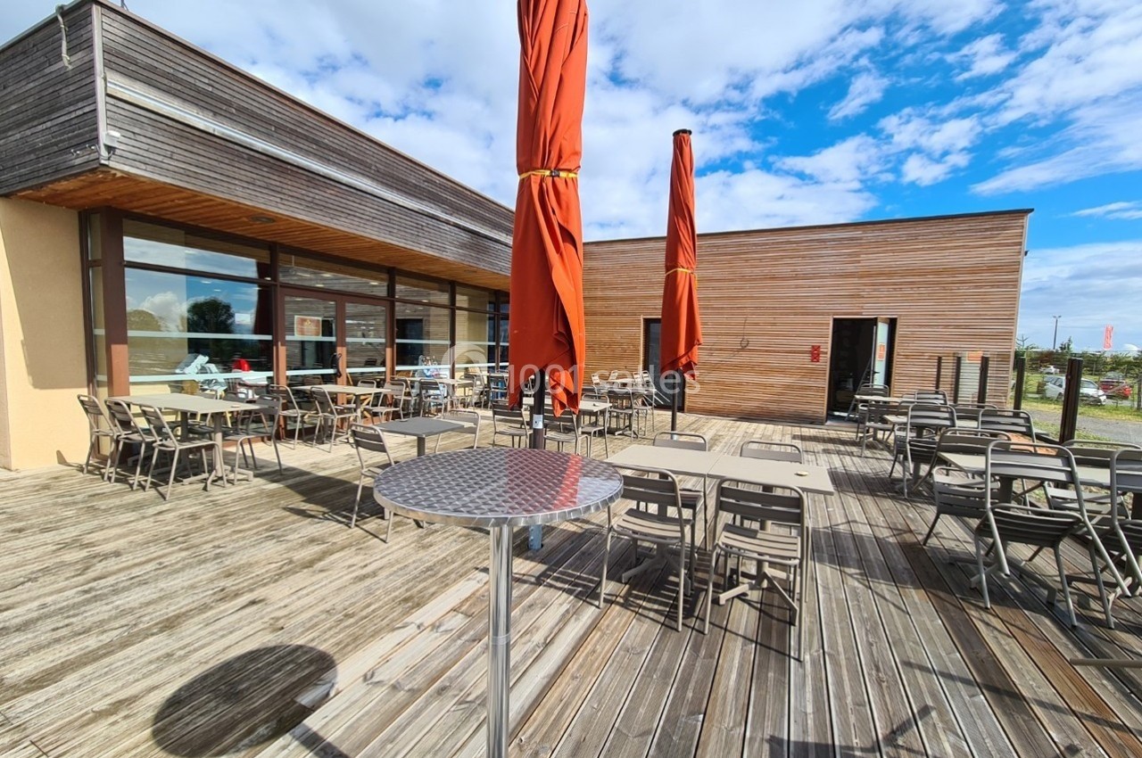 Terrasse en bois avec tables, chaises et parasols rouges, devant un bâtiment moderne sous un ciel partiellement nuageux.