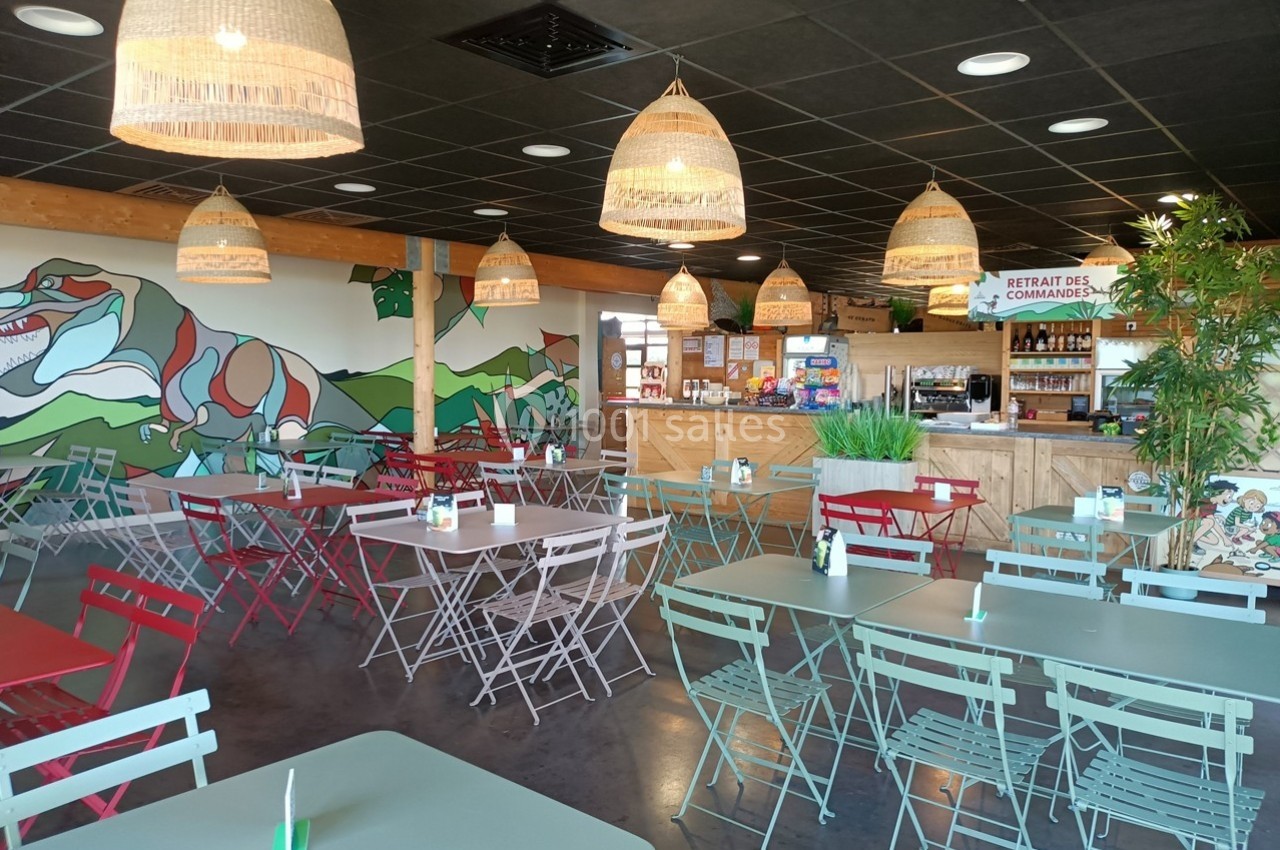 Salle de restaurant lumineuse avec tables et chaises colorées, décor mural végétal et comptoir de commande au fond.