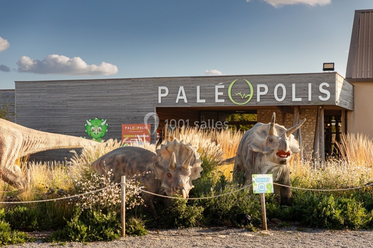 Reproduction de dinosaures devant l'entrée d'un parc nommé ’Paléopolis’, entourées de végétation.