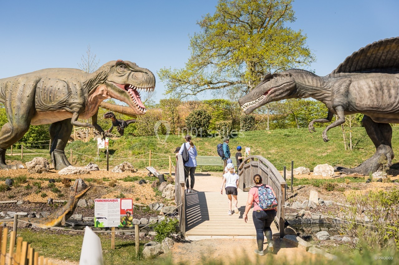 Des visiteurs traversent un pont dans un parc avec des statues grandeur nature de dinosaures sous un ciel dégagé.