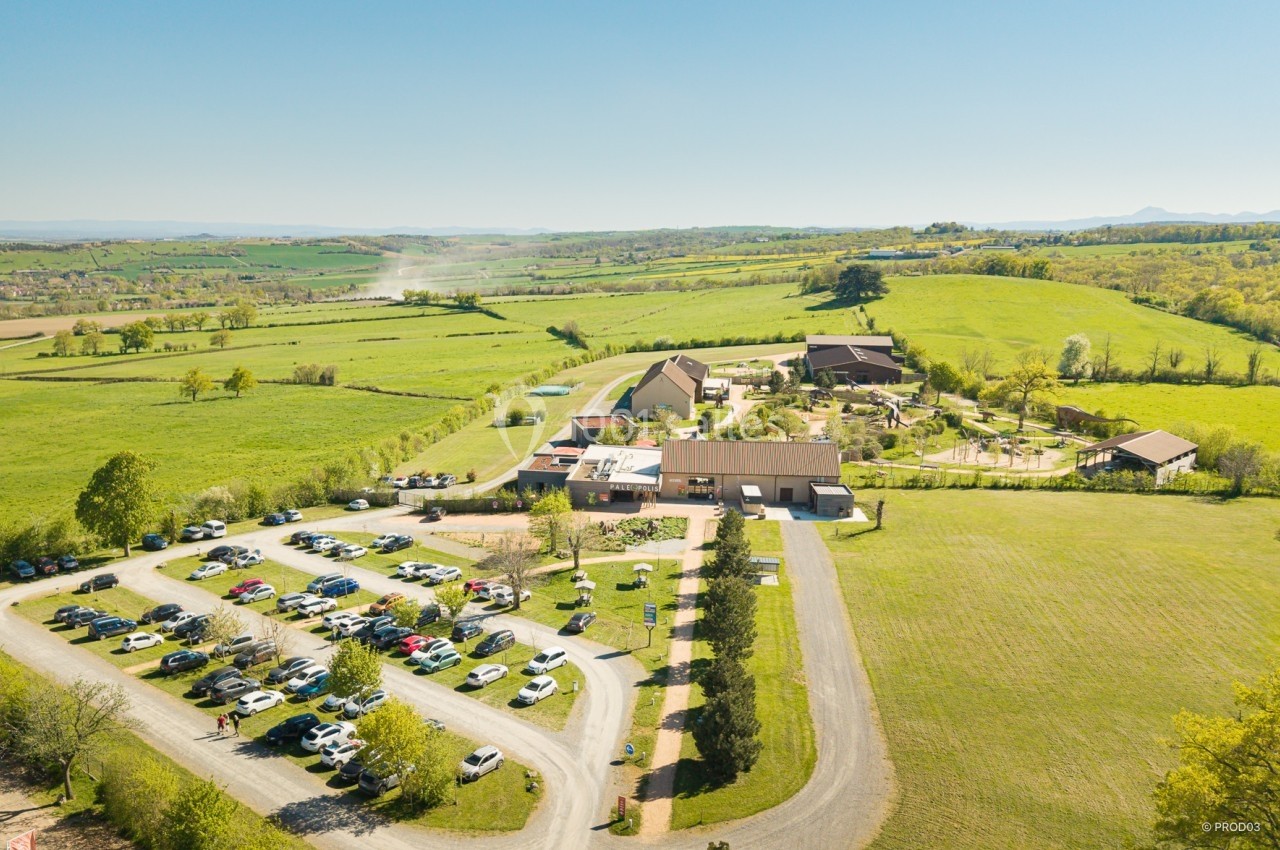 Vue aérienne d'un complexe rural avec bâtiments, parking rempli de voitures et paysage verdoyant environnant.