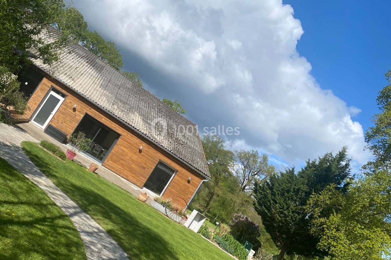 Maison en bois avec toit en tuiles, entourée d'un jardin verdoyant sous un ciel partiellement nuageux.