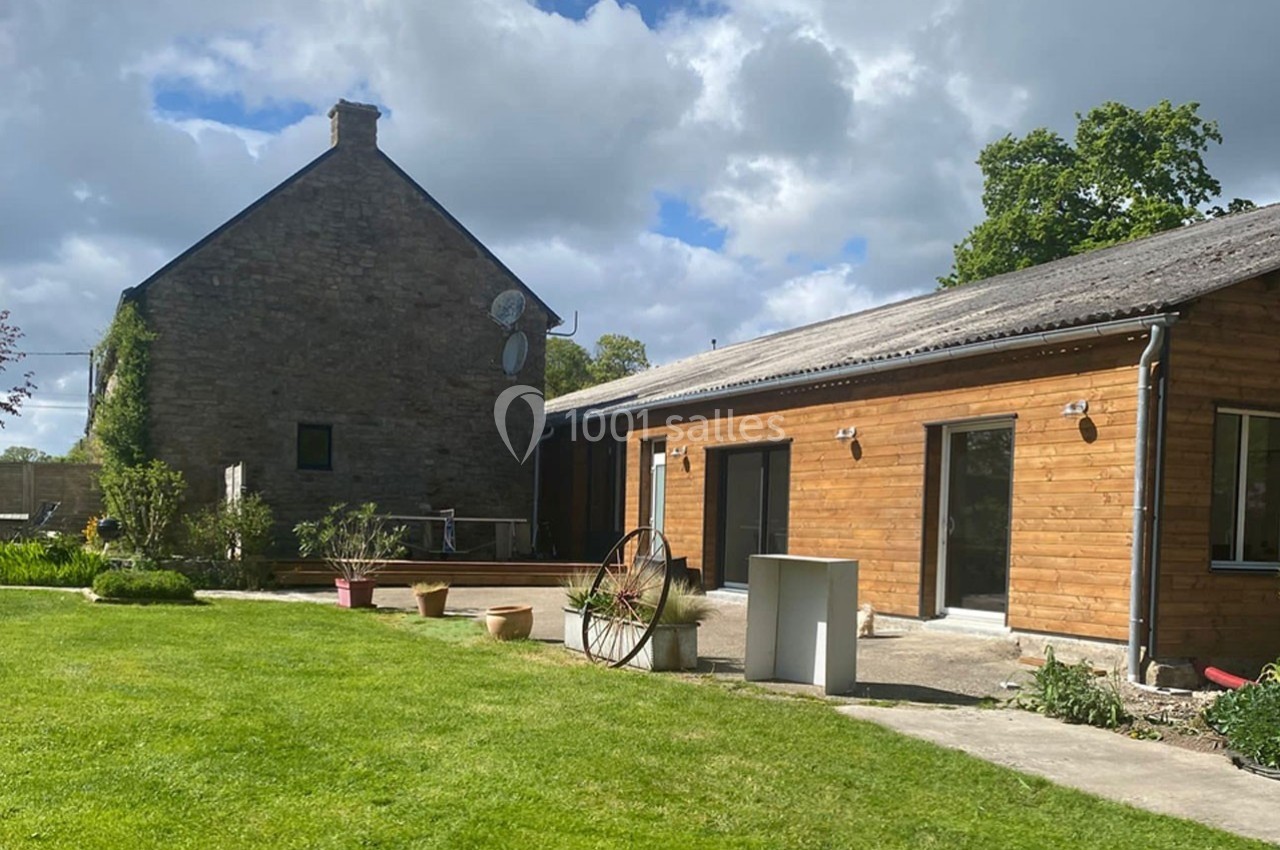 Maison en pierre et extension en bois avec jardin verdoyant sous un ciel partiellement nuageux.