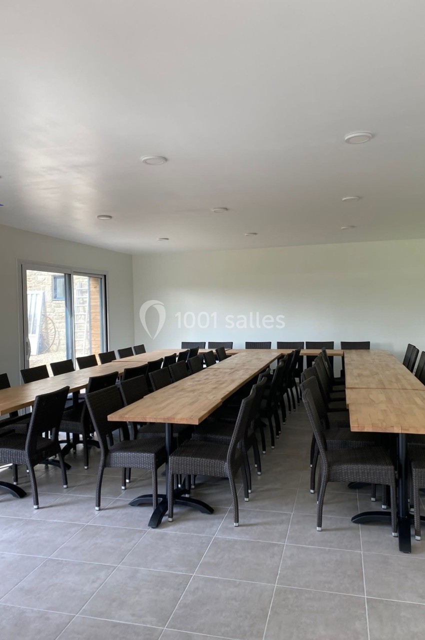Salle lumineuse avec deux longues tables en bois entourées de chaises noires, sol en carrelage gris.
