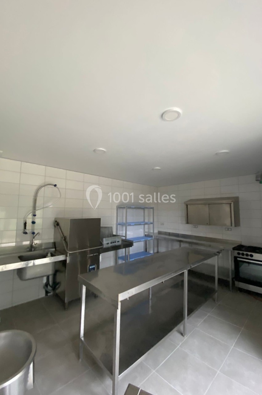 Cuisine professionnelle avec plan de travail en inox, évier, four, plaques de cuisson et murs carrelés blancs.