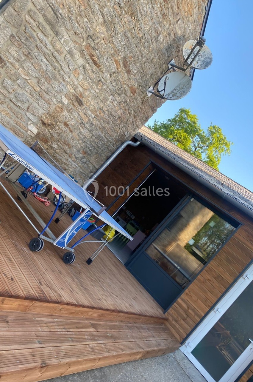 Table de ping-pong pliée sur une terrasse en bois, devant une maison en pierre avec des portes vitrées.
