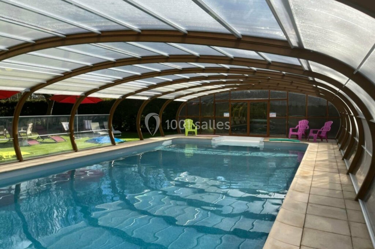 Piscine couverte avec structure en verre et métal, entourée de chaises colorées sur un sol en dalles.