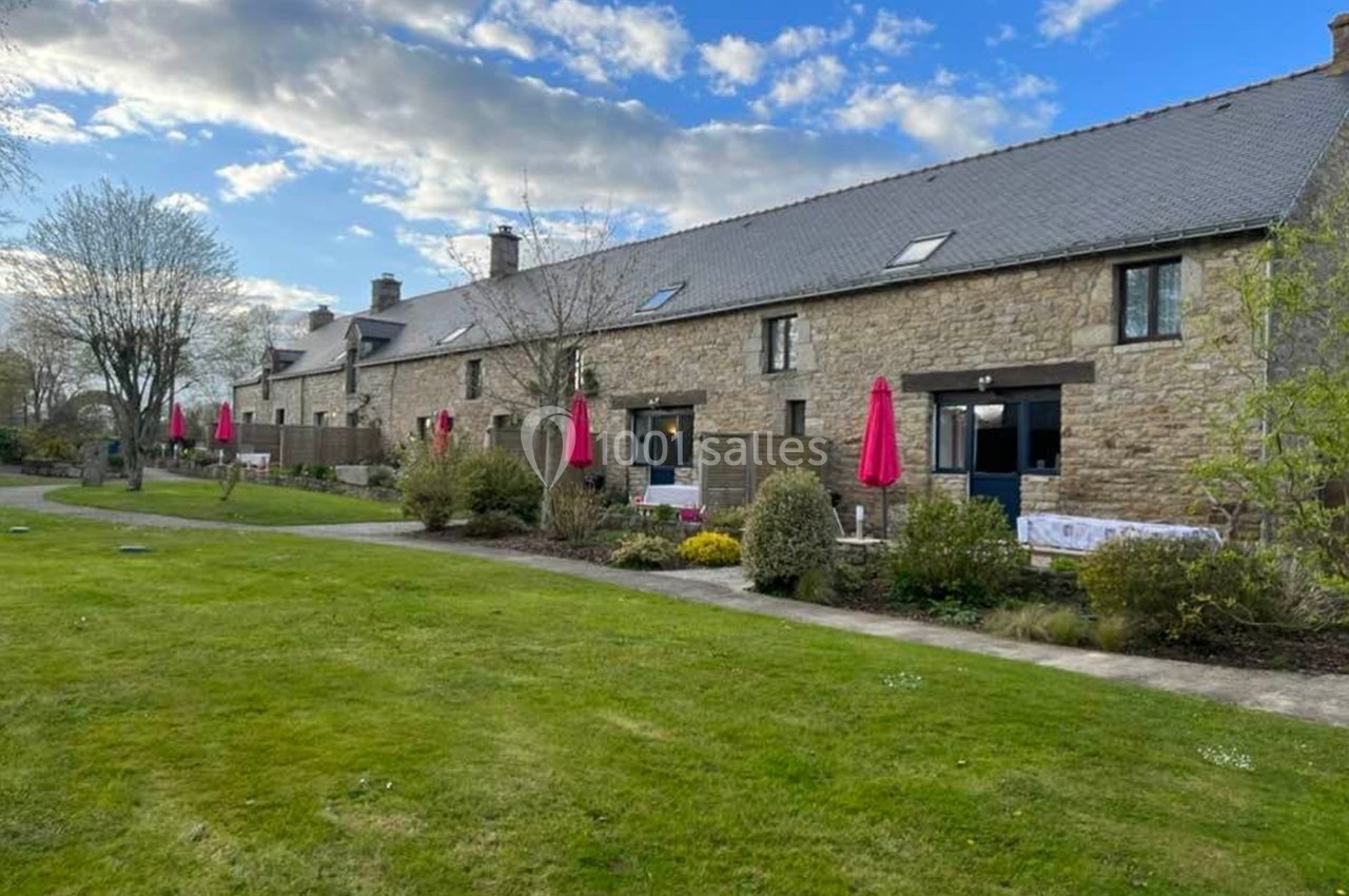 Long bâtiment en pierre avec des parasols rouges, entouré de pelouses et d'arbres sous un ciel partiellement nuageux.
