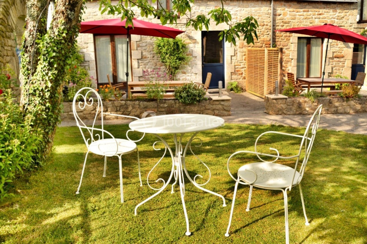 Mobilier de jardin en fer forgé blanc sur une pelouse, avec des parasols rouges et une façade en pierre en arrière-plan.