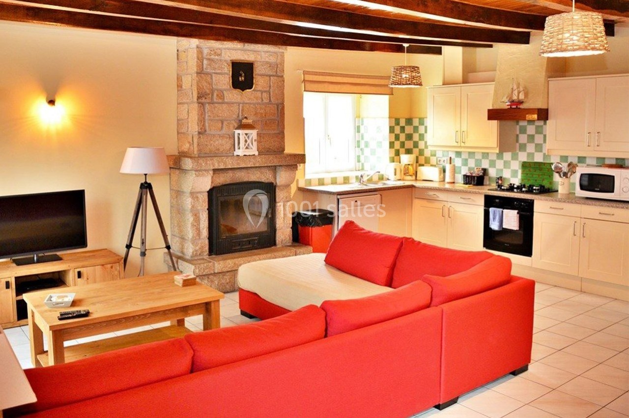 Salon avec canapé rouge en angle, cheminée en pierre, cuisine ouverte équipée et poutres apparentes au plafond.