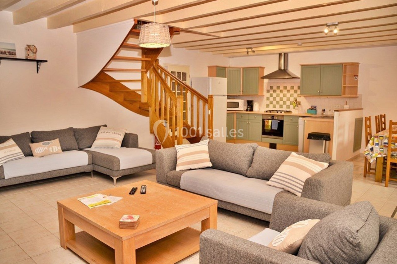 Salon lumineux avec canapés gris, table basse en bois, escalier en bois et cuisine ouverte équipée en arrière-plan.