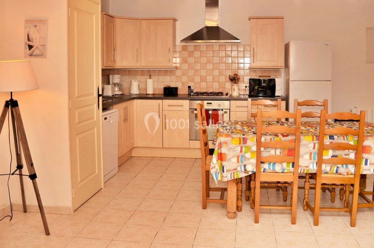 Cuisine équipée avec meubles en bois clair, table avec nappe colorée, réfrigérateur, lave-vaisselle et électroménagers.