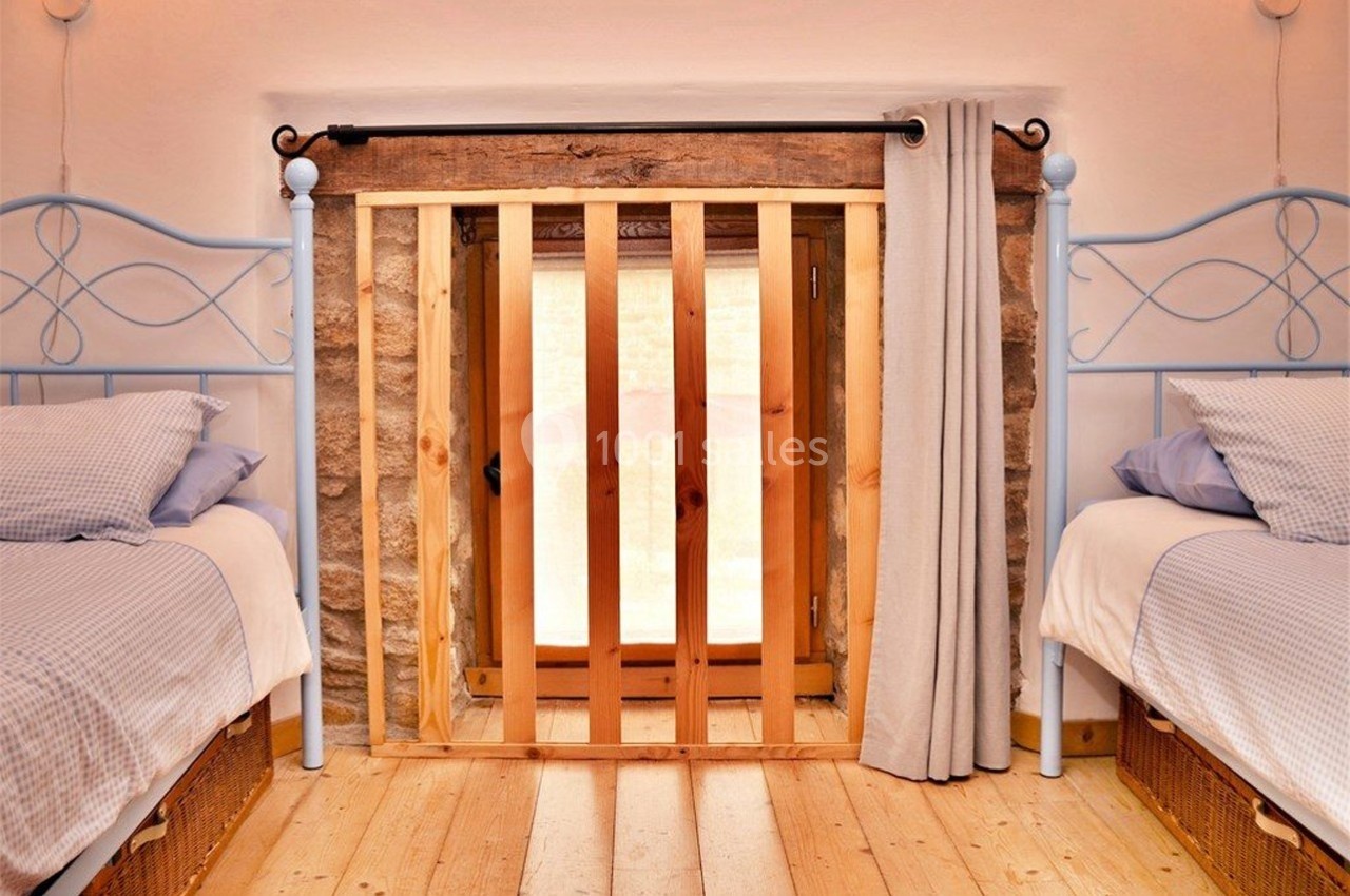 Deux lits simples avec linge bleu encadrent une petite porte en bois et pierre, partiellement couverte par un rideau beige.