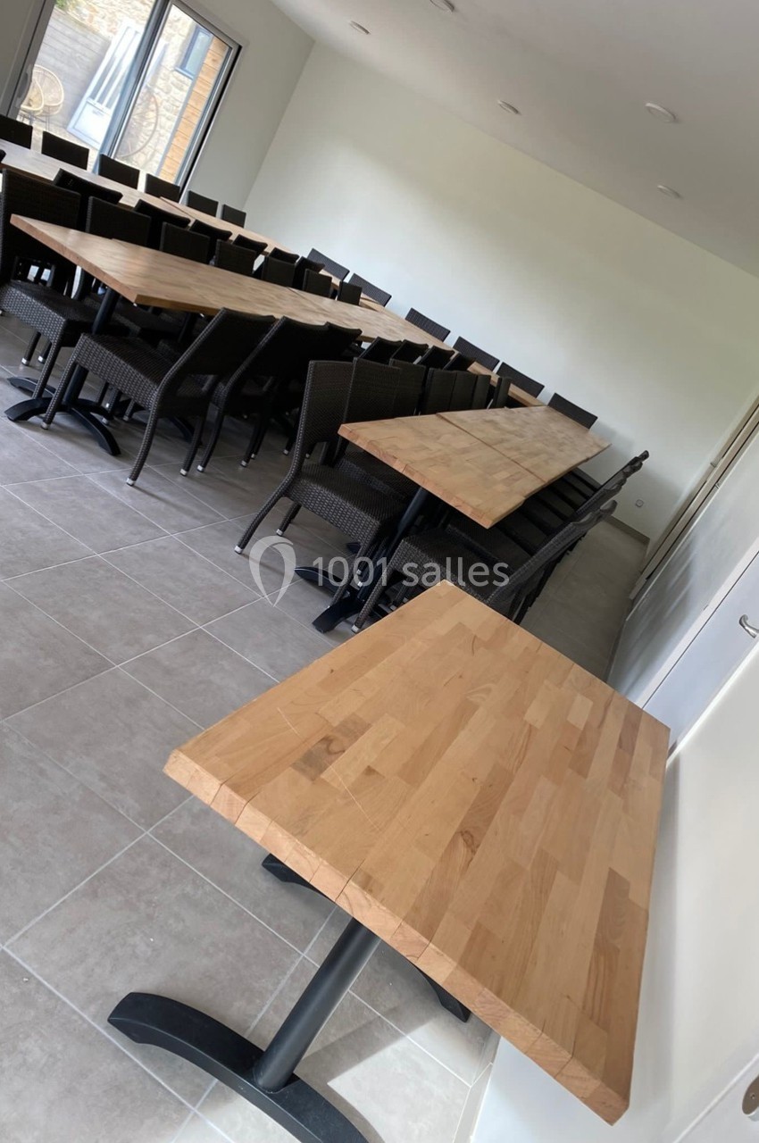 Salle lumineuse avec plusieurs tables en bois clair et chaises noires, disposées en rangées sur un sol carrelé.