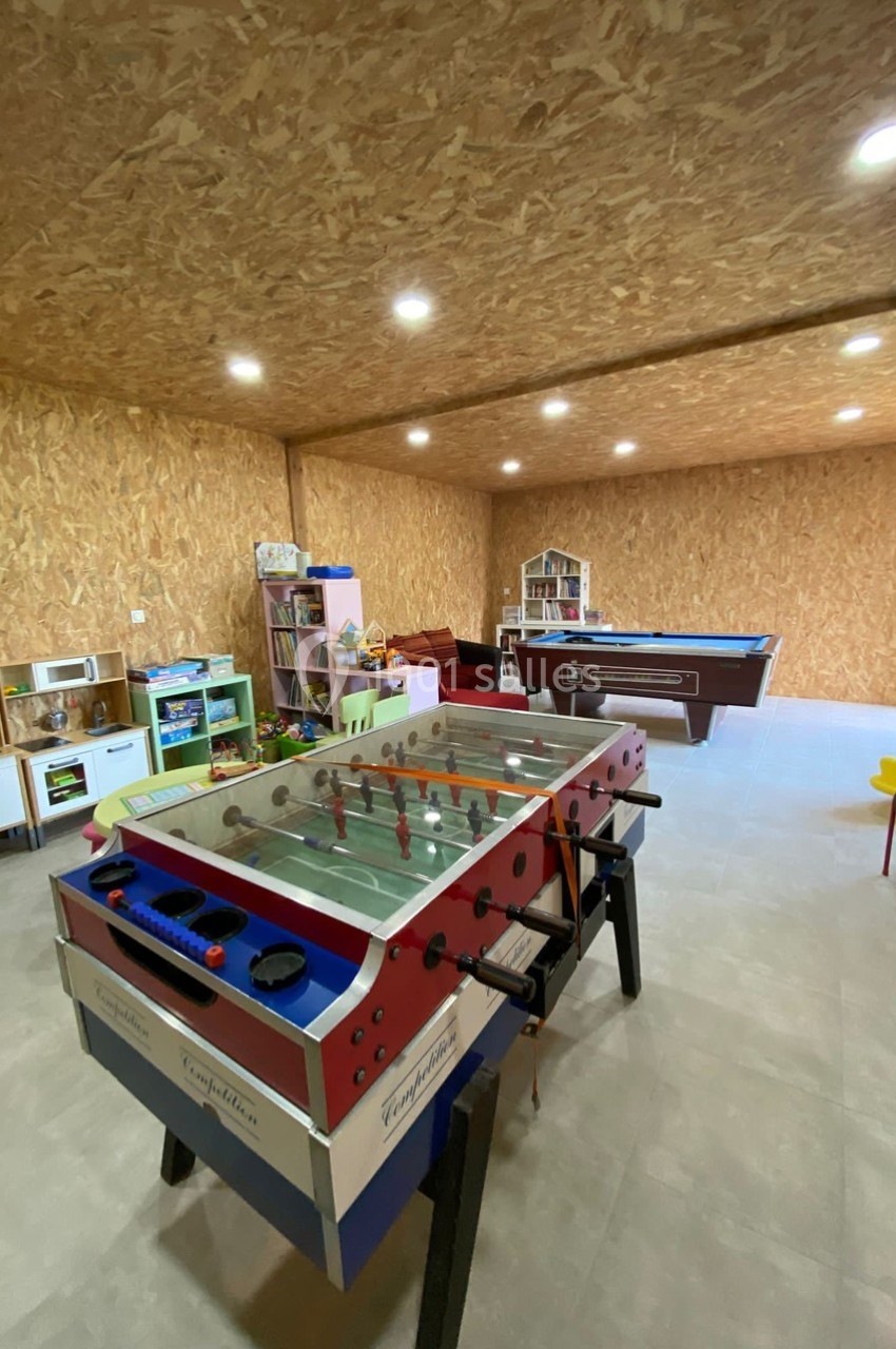 Salle de jeux avec baby-foot, billard, jouets pour enfants et mobilier coloré, dans un espace aux murs en bois.