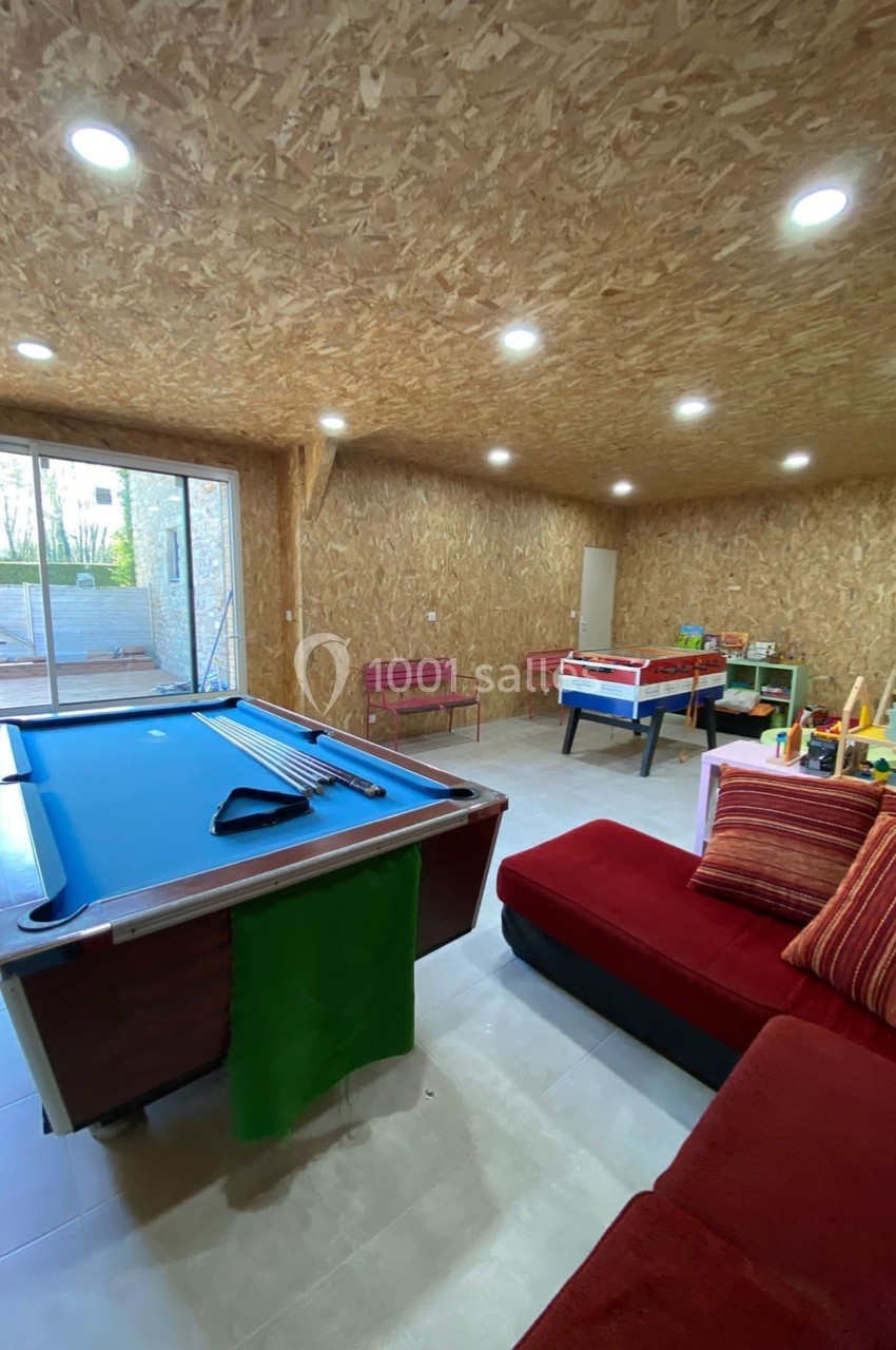 Salle de jeux avec billard, baby-foot, canapé rouge et grande baie vitrée donnant sur l'extérieur.