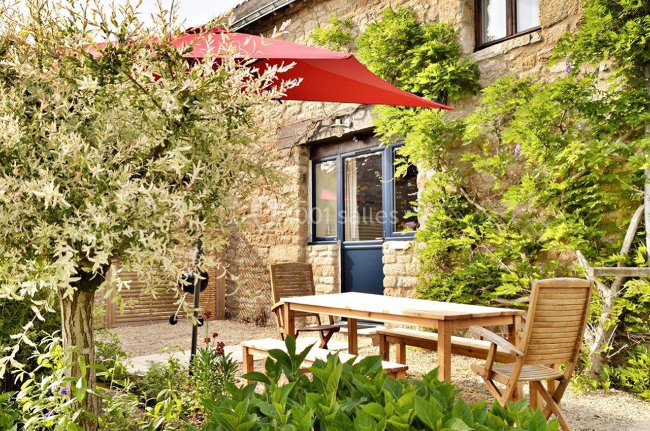 Table et chaises en bois sous un parasol rouge dans un jardin fleuri devant une maison en pierre.