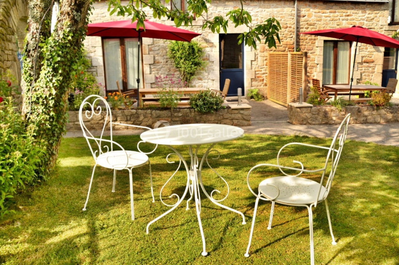 Mobilier de jardin en fer forgé blanc sur une pelouse, devant une maison en pierre avec parasols rouges.