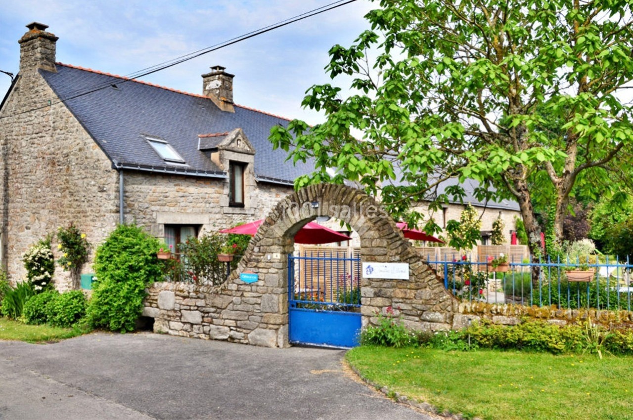 Maison en pierre avec toit en ardoise, portail en arche, jardin fleuri et terrasse avec parasols rouges.