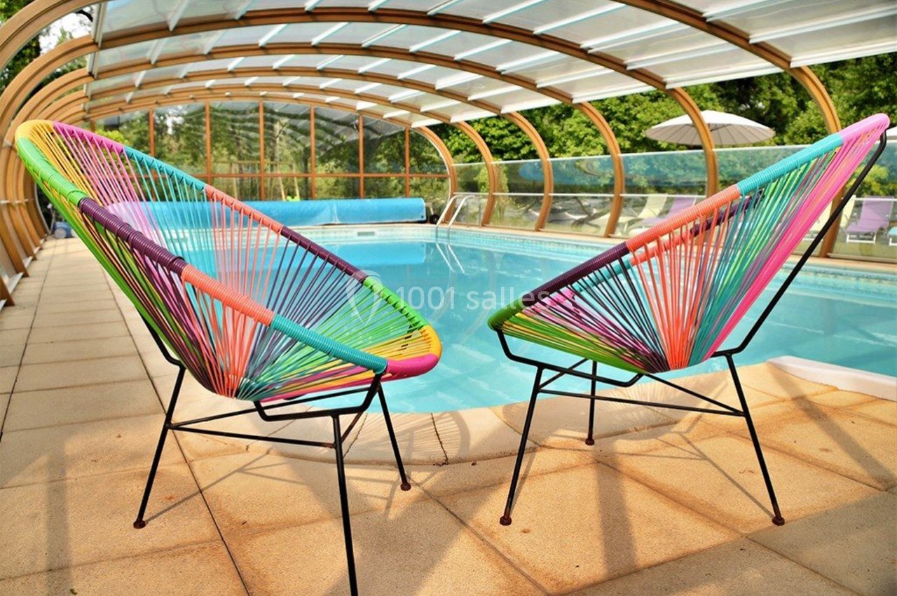 Deux fauteuils colorés en corde face à une piscine couverte par une structure en verre, entourée de végétation.