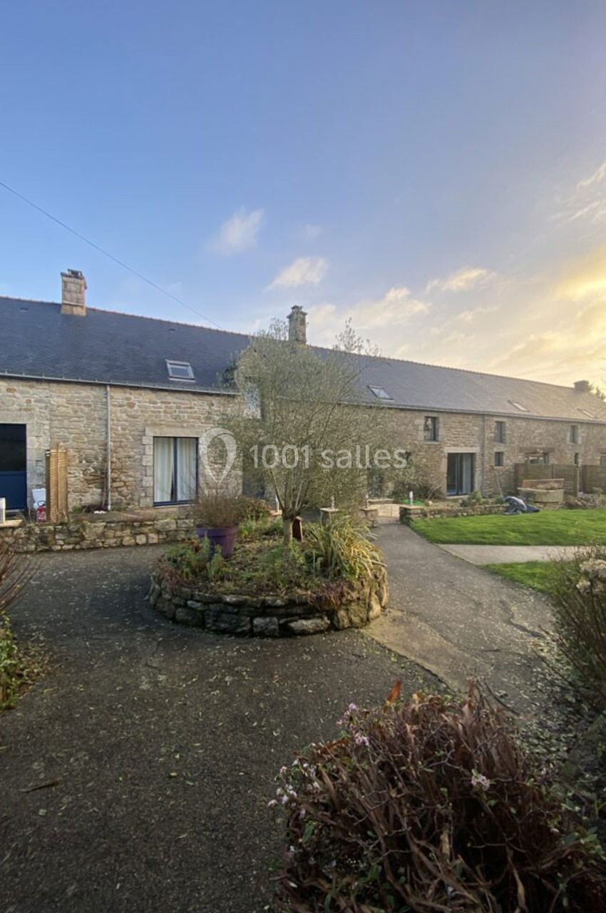 Cour d'une maison en pierre avec un arbre central, entourée de végétation et éclairée par une lumière douce.