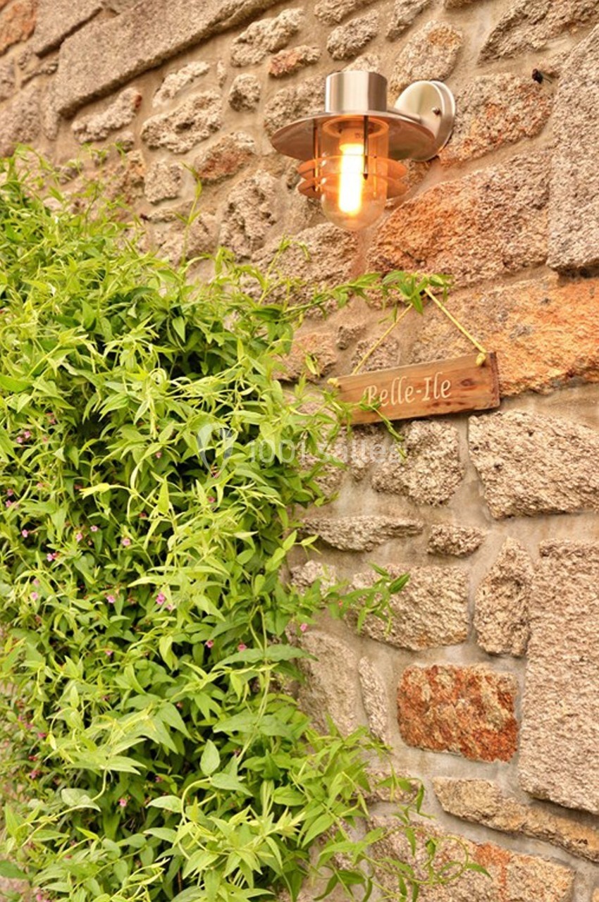 Applique murale allumée fixée sur un mur en pierre, entourée de verdure et d'une pancarte en bois marquée ’Belle-Île’.