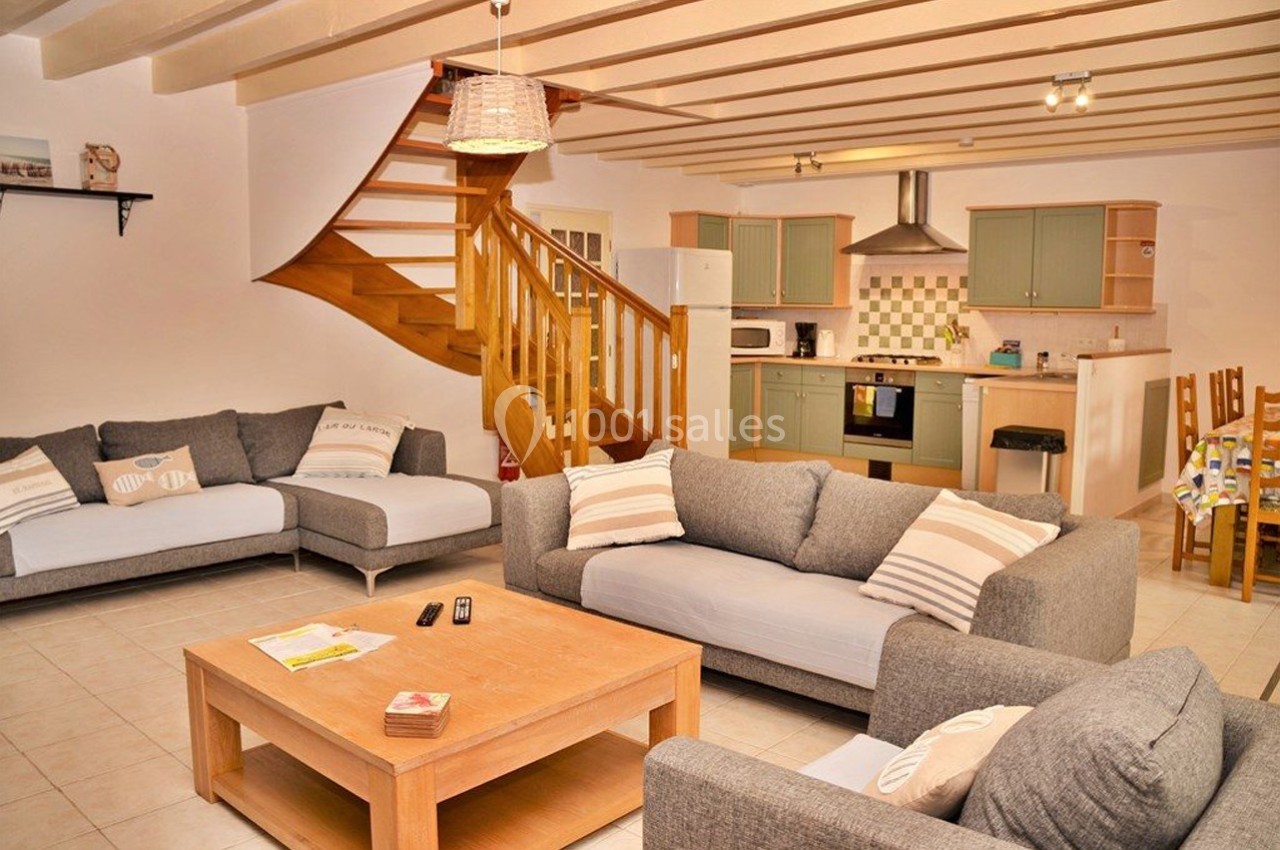 Salon lumineux avec canapés gris, table basse en bois, escalier en bois et cuisine ouverte équipée en arrière-plan.