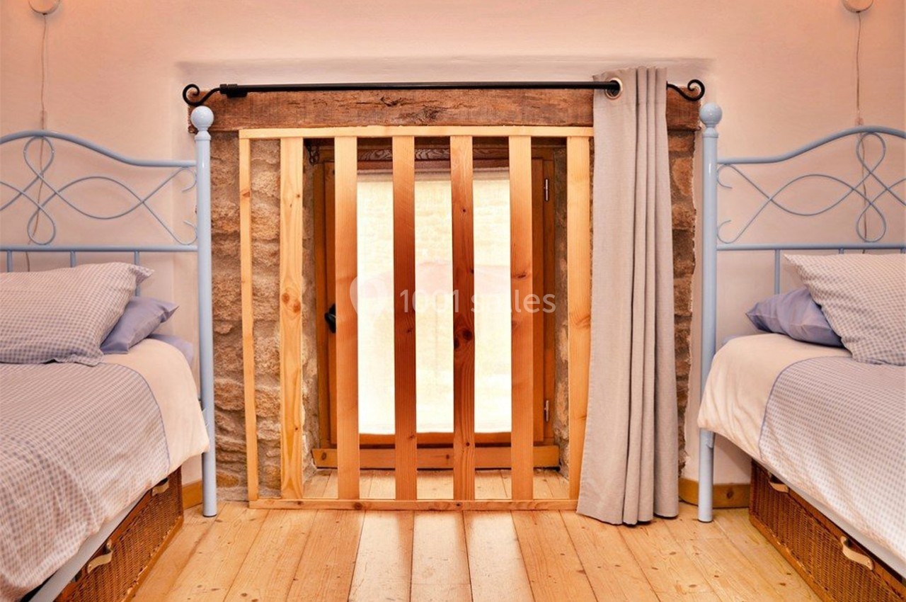 Deux lits simples avec draps bleus encadrent une petite porte en bois et pierre, partiellement couverte par un rideau beige.