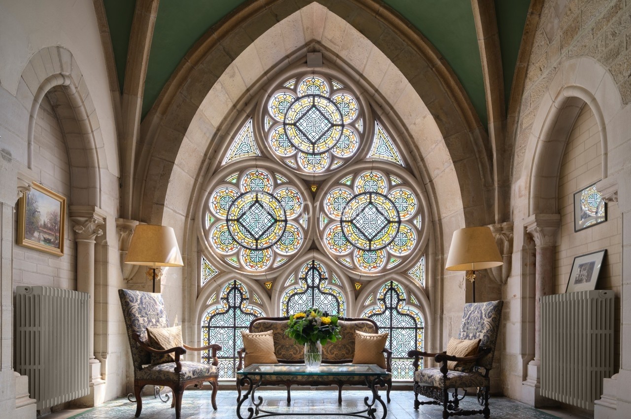Salle lumineuse avec grande fenêtre à vitraux colorés, deux fauteuils, une table basse et des lampes dans un décor ancien.