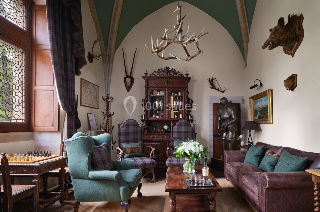Salon au style médiéval avec mobilier en bois, fauteuils, cheminée, armure, trophées de chasse et plafond voûté.