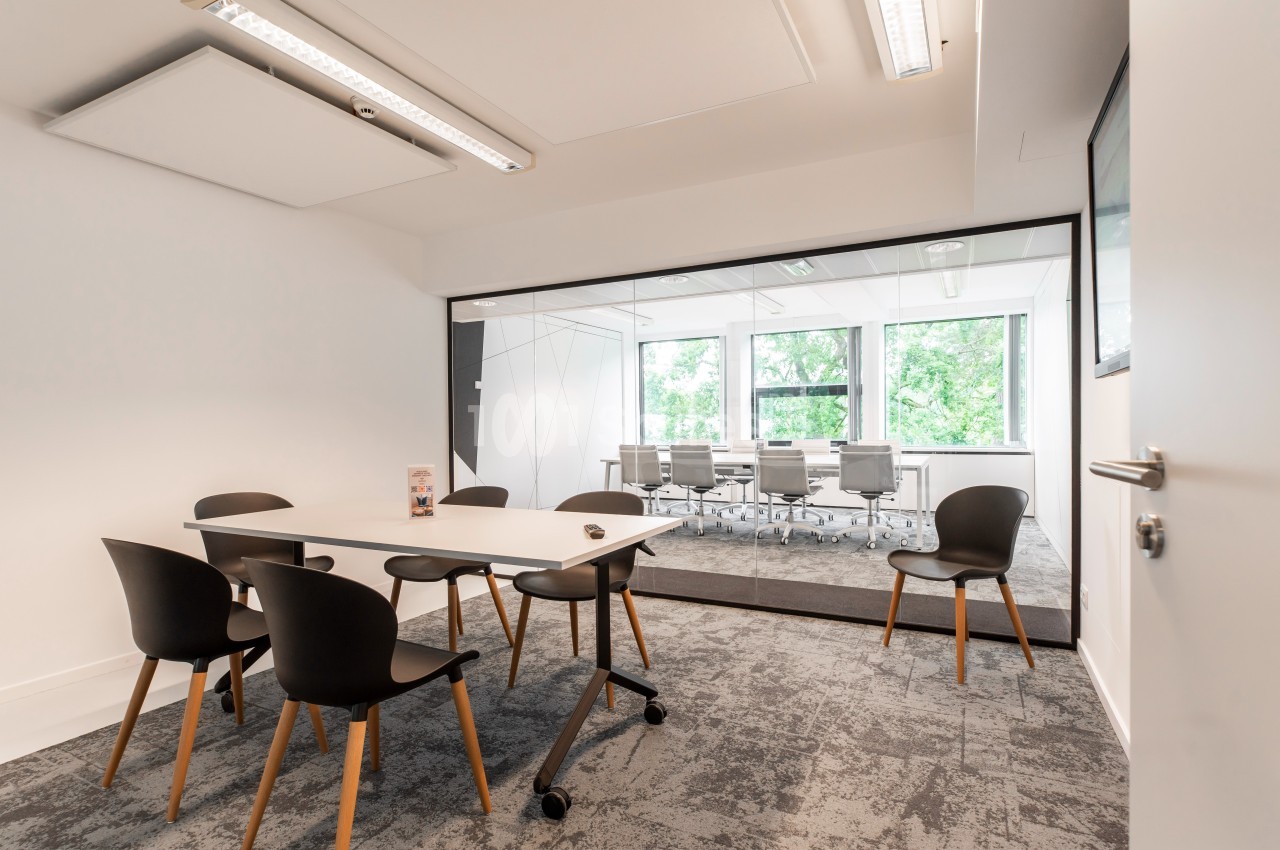 Salle de réunion moderne avec une table et des chaises noires au premier plan, et une autre salle vitrée en arrière-plan.