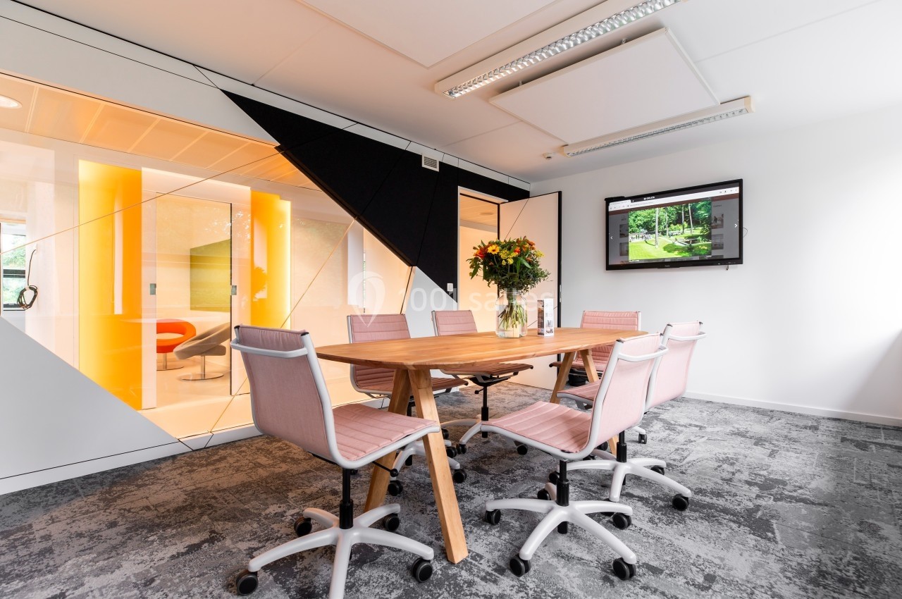 Salle de réunion moderne avec table en bois, chaises roses, écran mural et vase de fleurs, éclairée par une cloison vitrée.