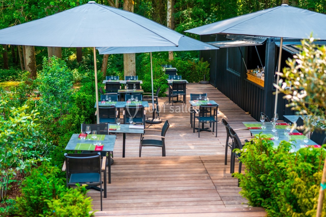 Terrasse en bois avec tables et chaises noires, entourée de verdure et équipée de grands parasols gris.