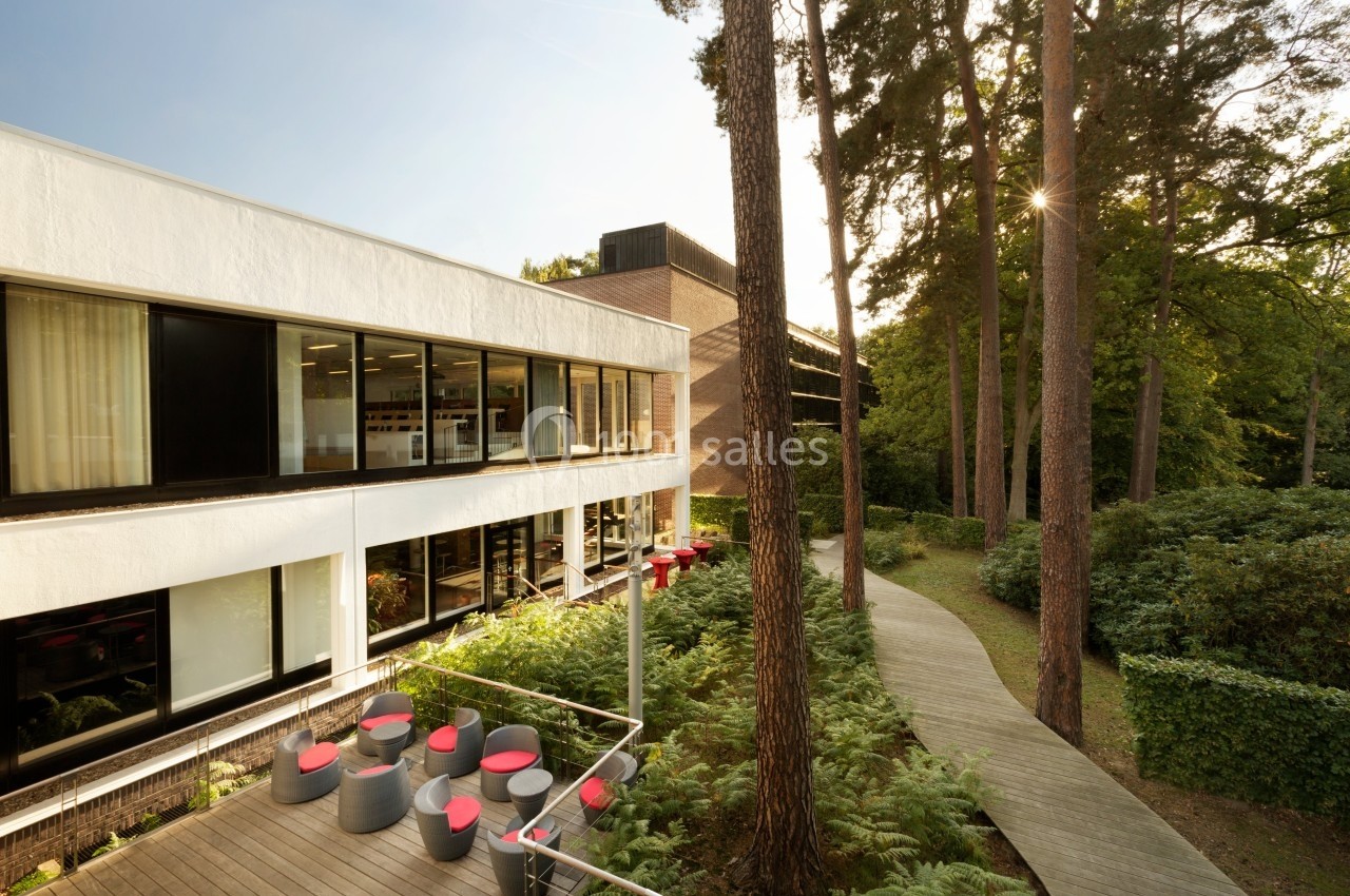 Bâtiment moderne entouré de végétation, avec une terrasse aménagée et un chemin en bois bordé d'arbres.