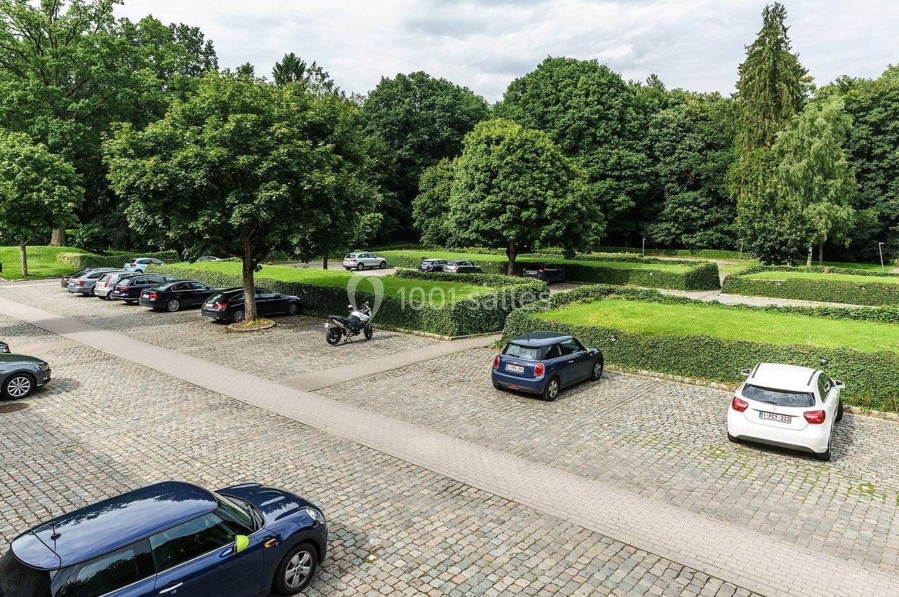 Parking extérieur pavé avec plusieurs voitures stationnées, entouré de haies et d'arbres verdoyants.