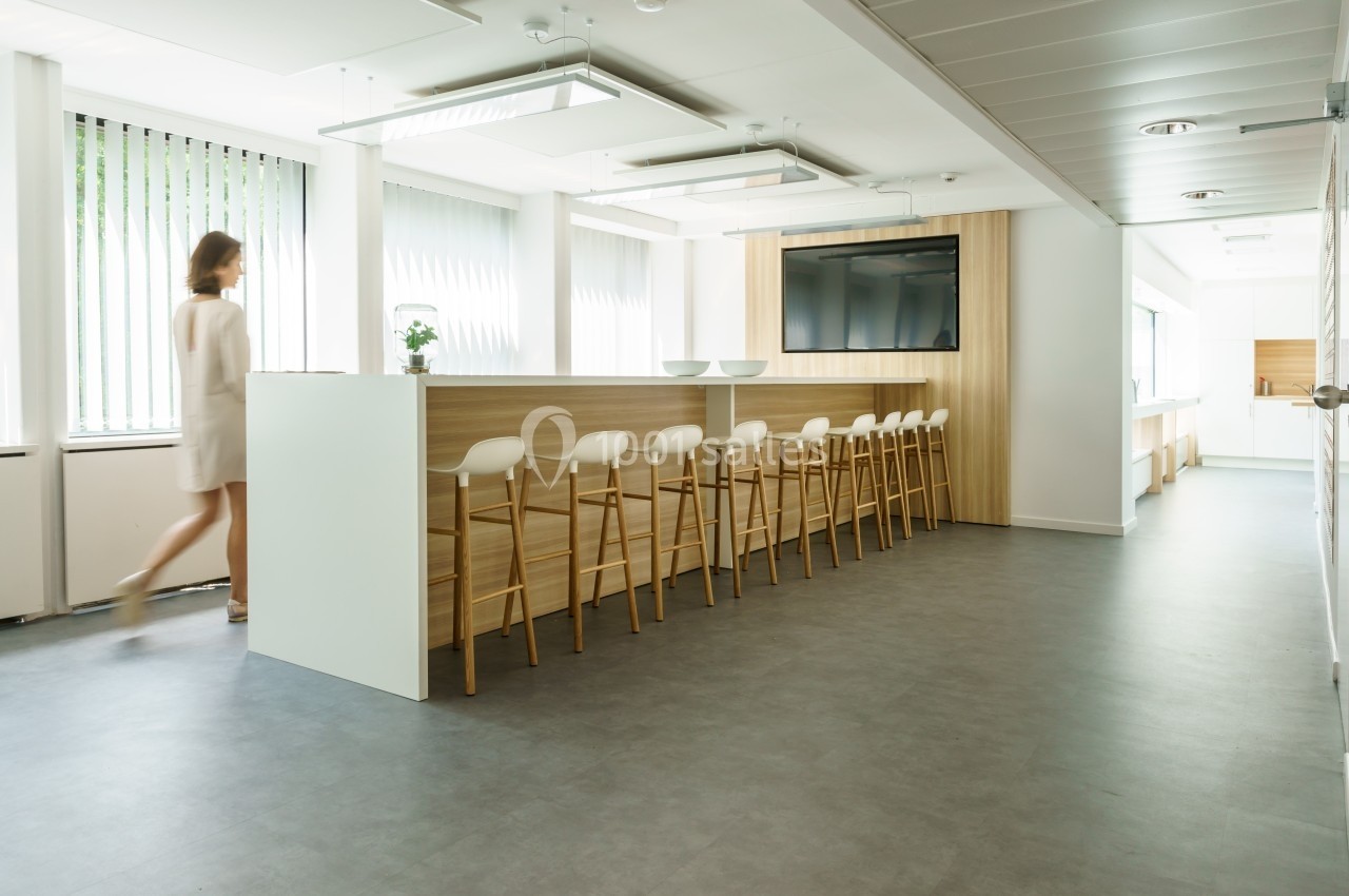 Une femme marche dans un espace lumineux avec un bar en bois, des tabourets hauts et des fenêtres à stores.