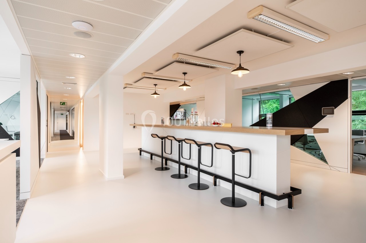 Espace lumineux avec un comptoir en bois, des tabourets noirs et un couloir menant à des bureaux vitrés.