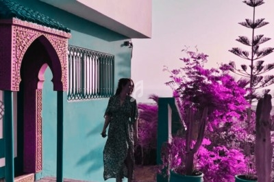 Femme marchant près d'une maison aux murs colorés, entourée de plantes en pots et d'arbres sous une lumière douce.