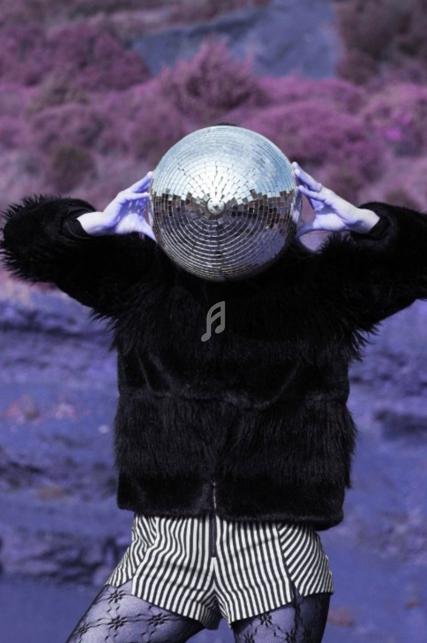 Personne portant une boule à facettes sur la tête, vêtue de noir et de rayures, dans un paysage violet surréaliste.