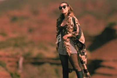 Femme debout sur un sol rouge, portant des lunettes de soleil et un manteau, avec un paysage flou en arrière-plan.