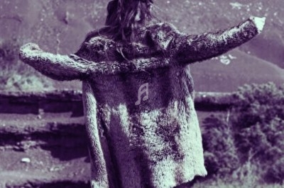 Femme de dos portant un manteau en fourrure, bras écartés, devant un paysage naturel avec collines.