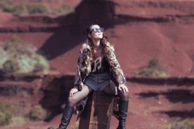 Femme assise sur une malle en plein air, entourée d'un paysage aride aux tons rouges et de formations rocheuses.