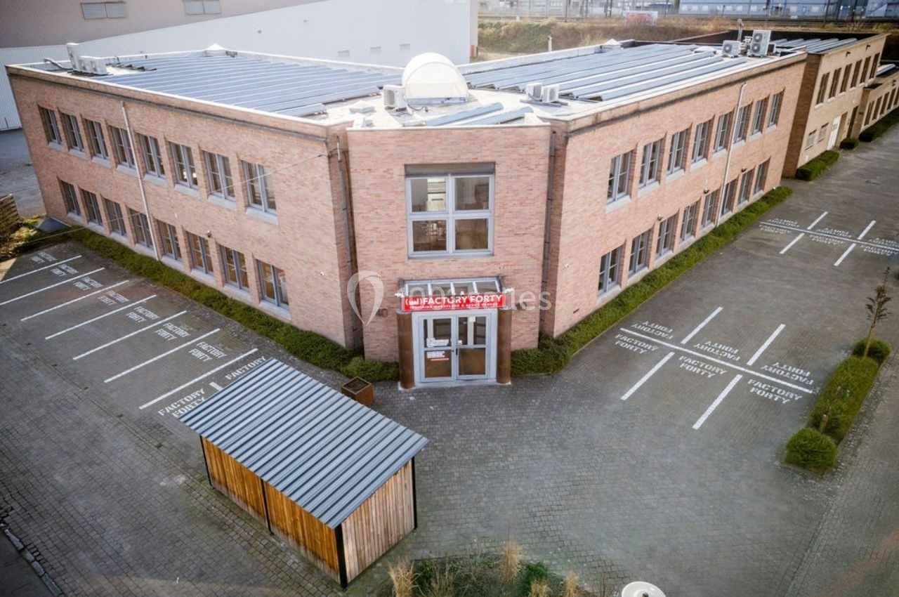 Bâtiment en briques à deux étages avec un parking adjacent et un abri en bois au premier plan.