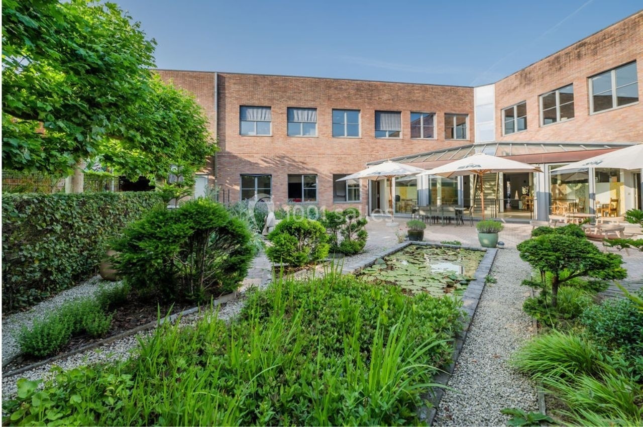 Cour intérieure avec jardin paysager, bassin de nénuphars, arbustes taillés et terrasse avec parasols devant un bâtiment en…