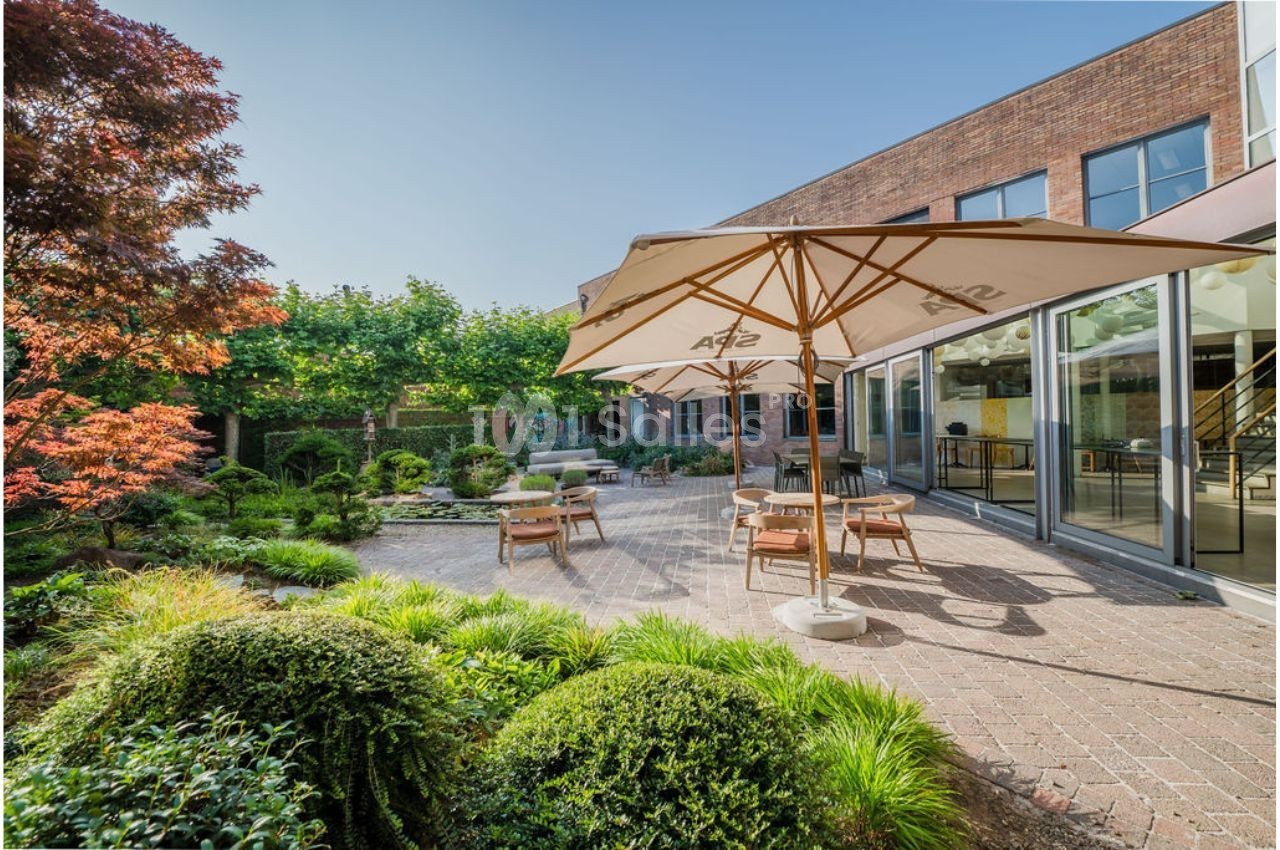 Terrasse ensoleillée avec tables, chaises et parasols, entourée de verdure et jouxtant un bâtiment en briques.