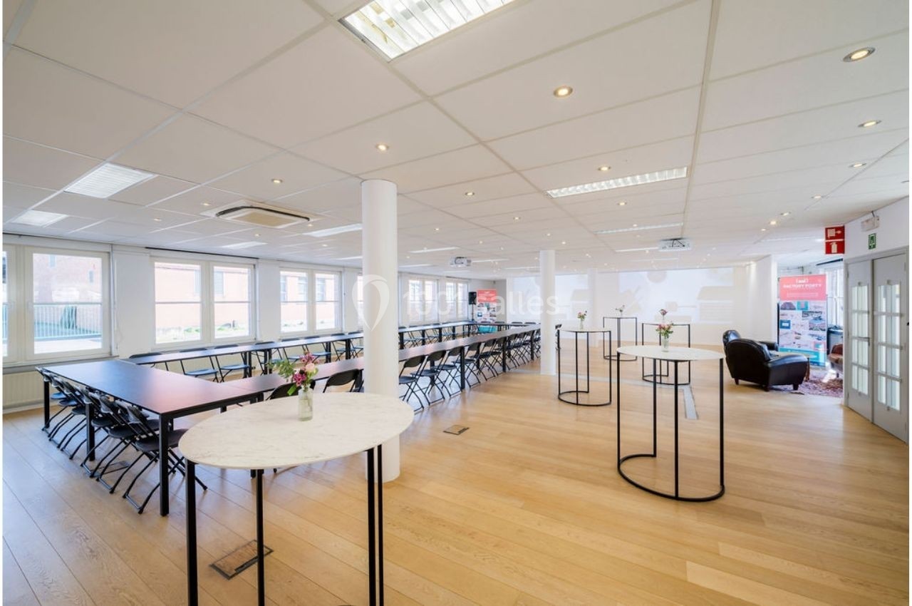 Salle lumineuse avec grandes fenêtres, tables alignées, chaises pliantes et petites tables rondes sur un sol en bois clair.