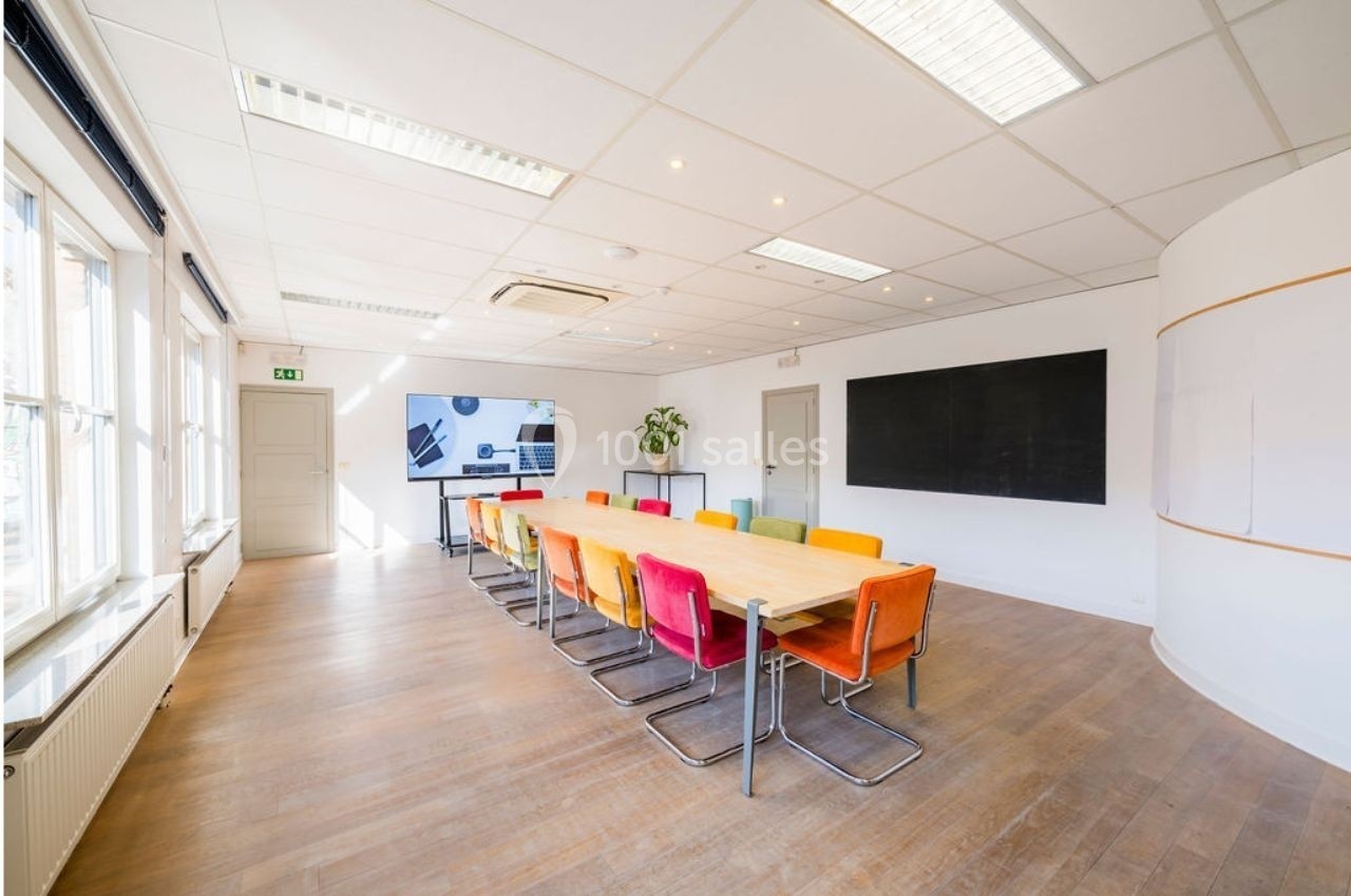 Salle de réunion lumineuse avec une grande table en bois entourée de chaises colorées et un écran mural.