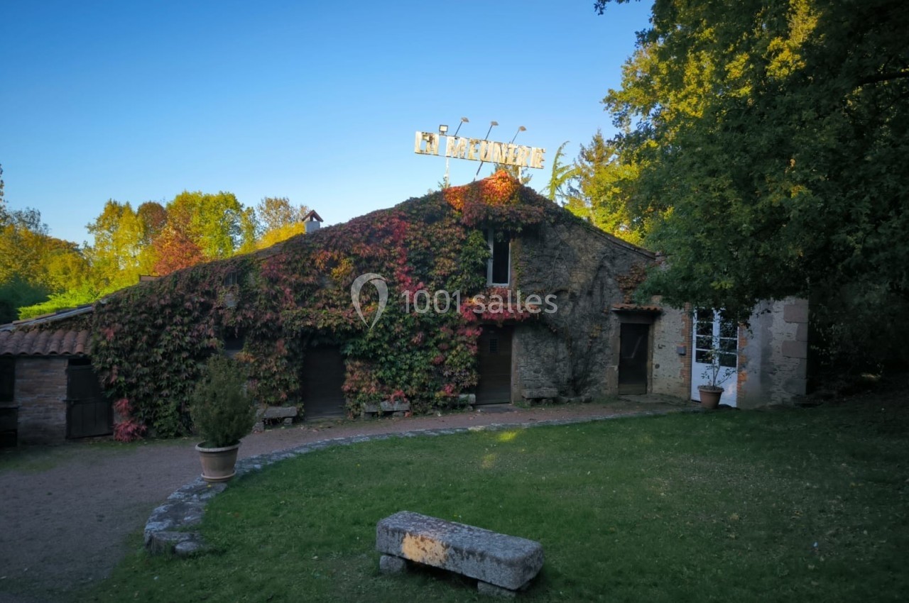 Bâtiment en pierre couvert de végétation avec une enseigne lumineuse, entouré d'arbres et d'un banc en pierre au premier…