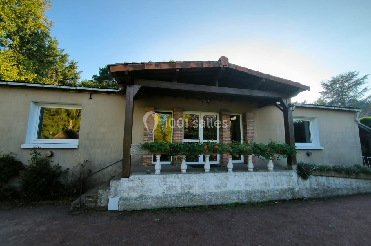 Façade d'une maison avec terrasse couverte, jardinières fleuries et murs clairs, entourée de végétation.