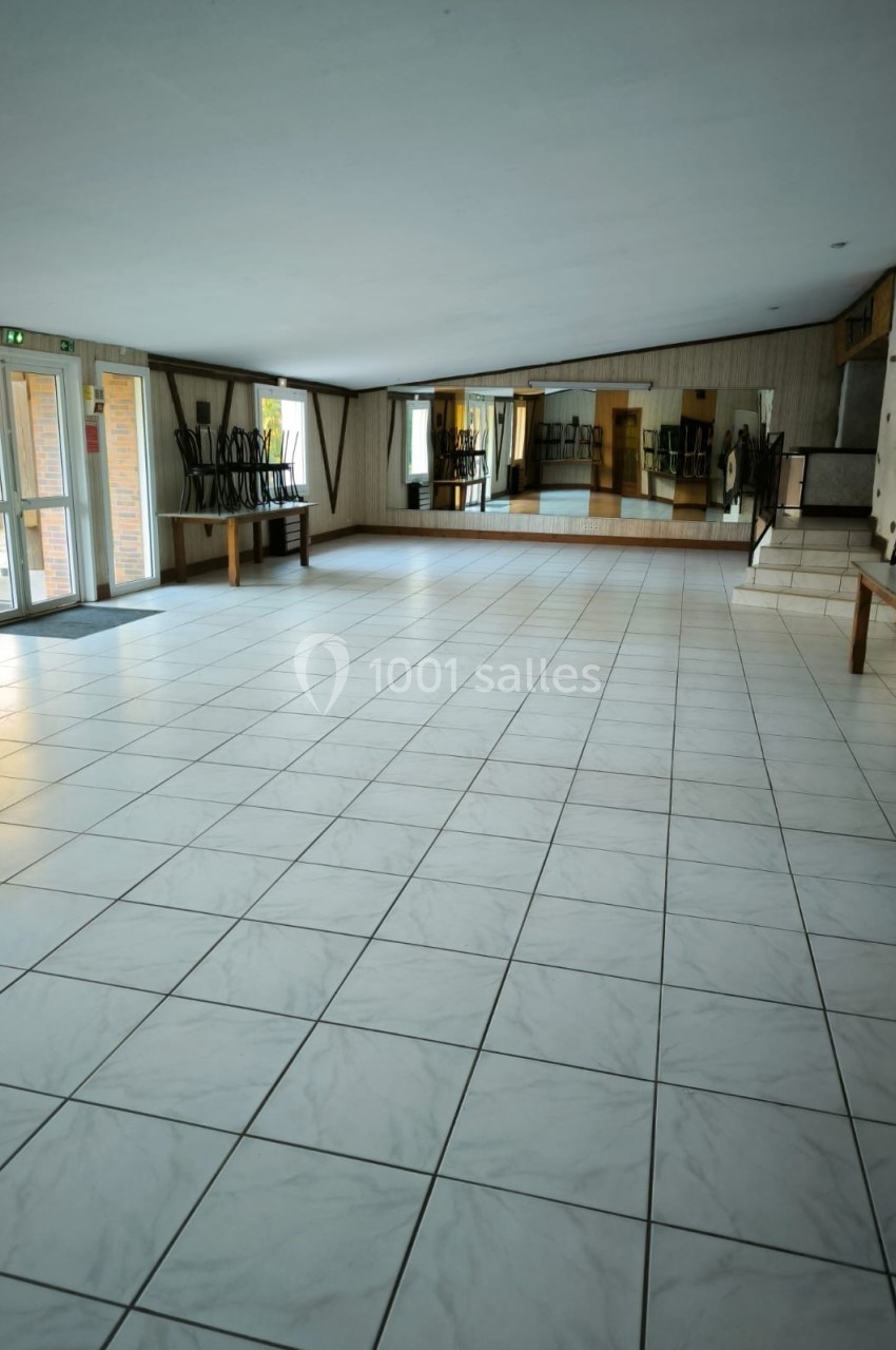 Salle vide avec carrelage blanc, chaises empilées sur des tables en bois, miroirs au mur et lumière naturelle.